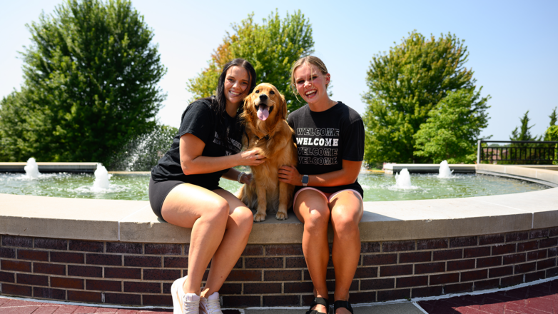 Two female student sitting by fountain with golden retriever