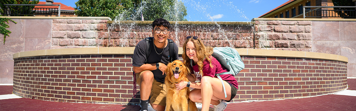 Two Students Sitting In Front Of Fountain With Golden Retriever