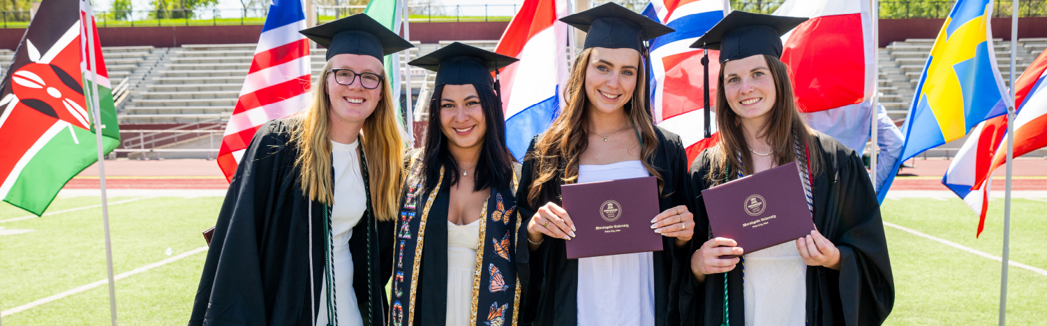 four graduates standing in front of international flags at commencement